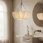 A modern bathroom scene featuring a hanging pendant light with a pleated beige shade, a large round wall mirror, a white vessel sink, and folded towels.