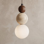 A modern three-sphere pendant light hanging against a textured beige plaster wall. The light features a dark brown top sphere, a speckled cream middle sphere, and a glowing warm white bottom globe shade.