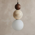 A modern three-sphere pendant light hanging against a textured beige plaster wall. The light features a dark brown matte sphere on top, a natural beige travertine stone sphere in the middle, and a frosted white glass sphere on the bottom.