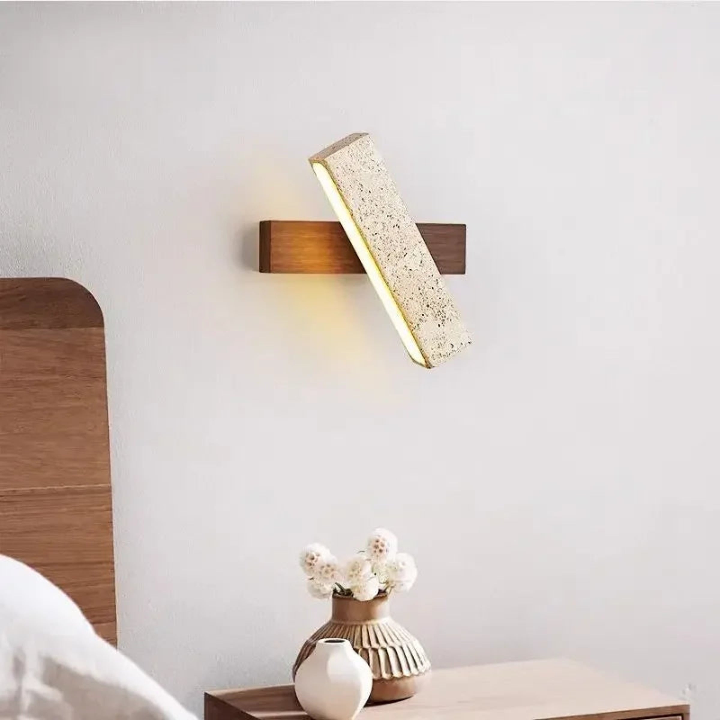 Modern wall lamp with wooden finish and textured shade on a white wall above a wooden nightstand with a vase.