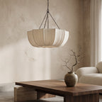 A modern pendant light with a pleated, drum-like shade hangs in a minimalist living room with textured beige walls, a wooden coffee table, and a cream sofa.
