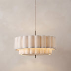 A modern drum-shaped pendant light with fluted, pleated shades in a warm cream or beige tone, suspended by a thin cord against a textured beige wall.