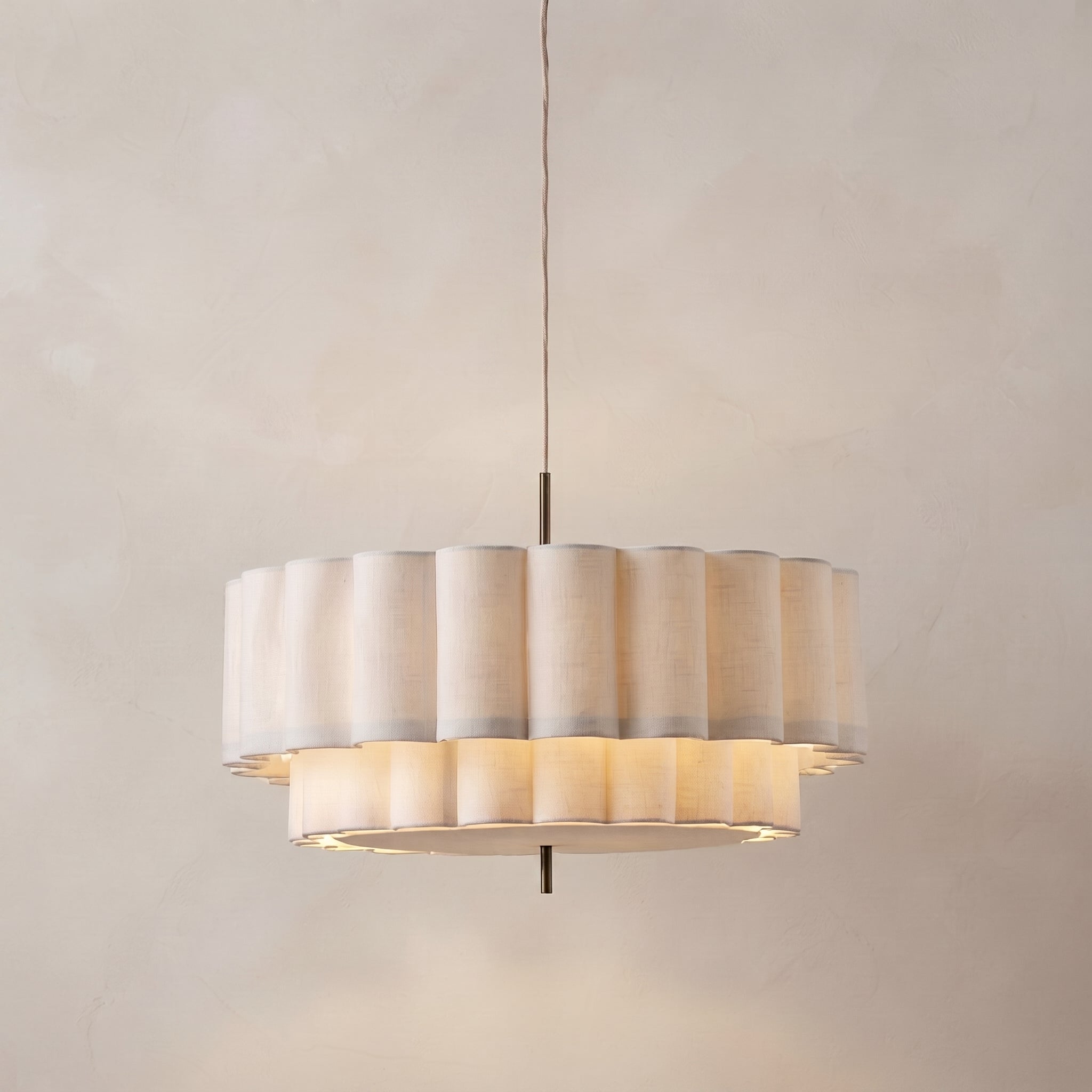 A modern drum-shaped pendant light with fluted, pleated shades in a warm cream or beige tone, suspended by a thin cord against a textured beige wall.