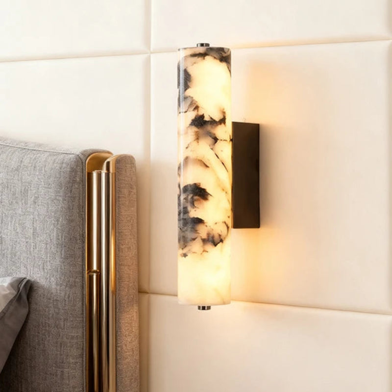 A modern wall-mounted sconce light fixture with a cylindrical marble-look shade featuring black and cream veining, illuminated with a warm glow. It is mounted on a cream-colored tiled wall next to a gray upholstered headboard with gold trim.