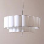 A modern circular pendant light with a white fluted fabric shade and a black hanging cord, shown against a plain beige wall.