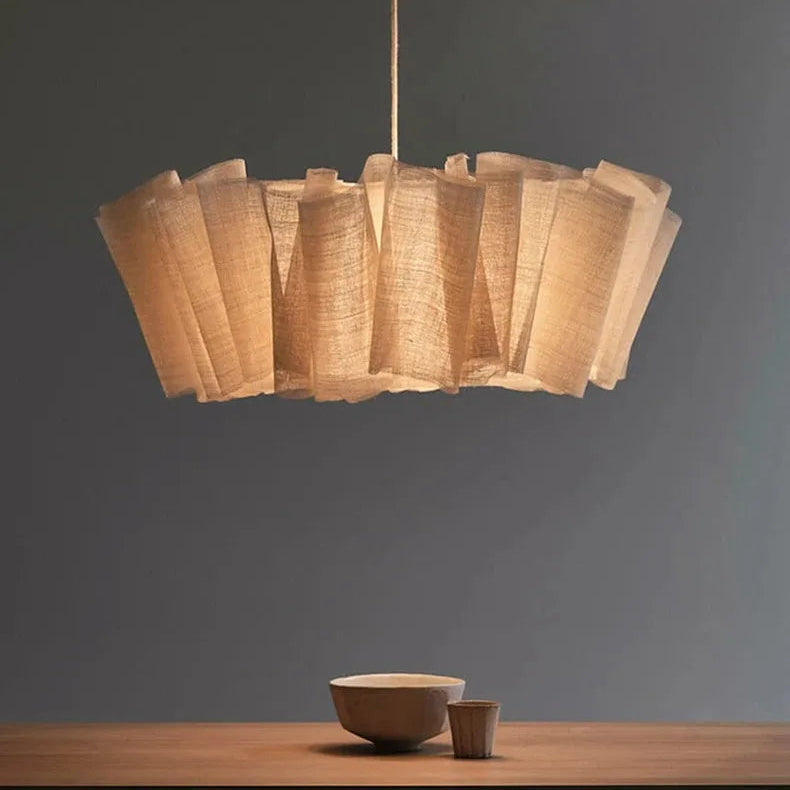 A modern pendant light with a tiered, ruffled shade made of natural beige woven material, suspended over a wooden table with a bowl and cup against a dark gray wall.