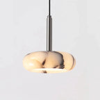 A modern pendant light with a rounded, disc-shaped translucent shade in a cream and black marbled pattern, suspended by a slim vertical metallic rod against a light gray background.