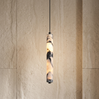 A modern cylindrical pendant light with a marbled stone or resin shade glowing warm white, suspended by a thin black cord against a textured beige stone wall.