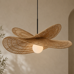 A modern pendant light with a large, organic-shaped woven rattan shade in a natural tan color, suspended by a thin black cord with a glowing white globe bulb inside, set against a warm beige textured wall.