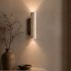 A modern cylindrical wall sconce with a textured beige stone or marble-like finish, mounted on a warm beige wall and casting a soft downward light. A ceramic vase with dried branches and a small bowl sit on a tabletop below.