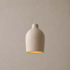 A modern pendant light with a textured, matte beige ceramic or stone-like shade, suspended by a thin white cord against a neutral, warm beige background.