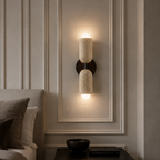A modern wall-mounted sconce with two cylindrical stone shades in a warm beige tone, mounted on a dark metal bracket and illuminated against a beige paneled wall in a bedroom setting.