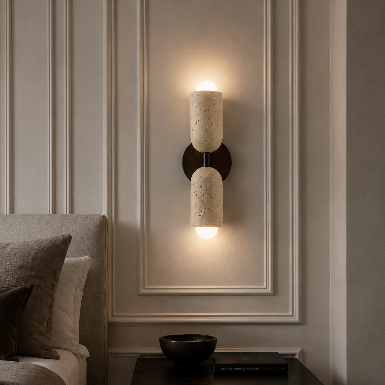 A modern wall-mounted sconce with two cylindrical stone shades in a warm beige tone, mounted on a dark metal bracket and illuminated against a beige paneled wall in a bedroom setting.