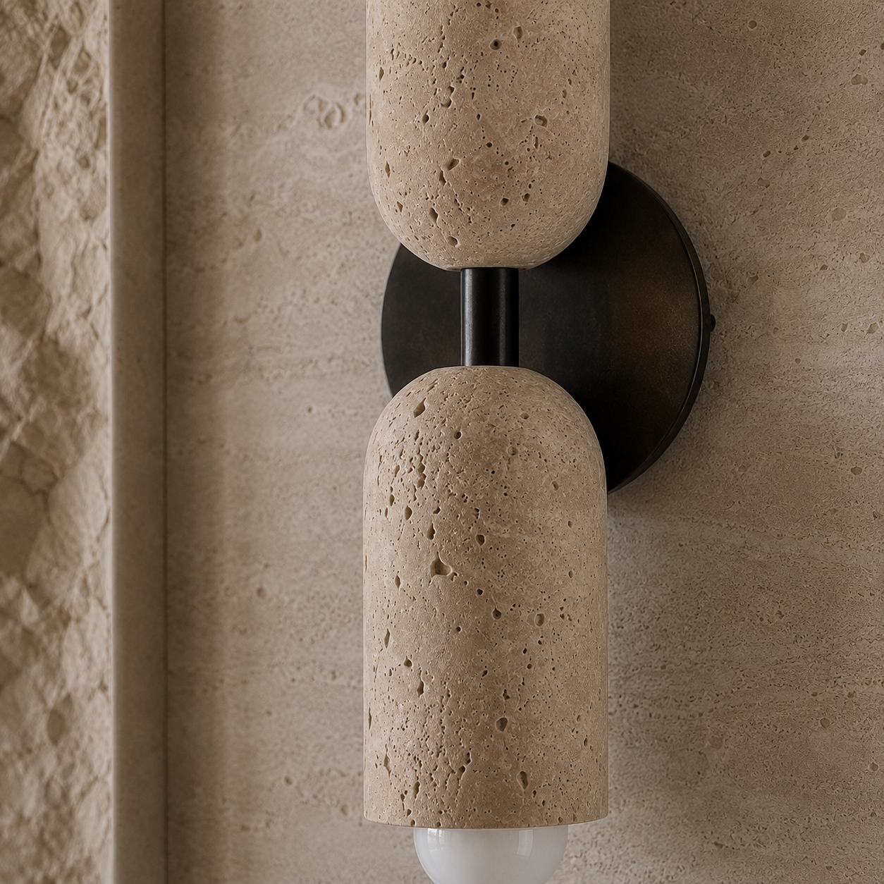 A modern wall-mounted sconce with two cylindrical stone shades in a textured beige travertine finish, mounted on a dark metal circular backplate against a matching beige stone wall.