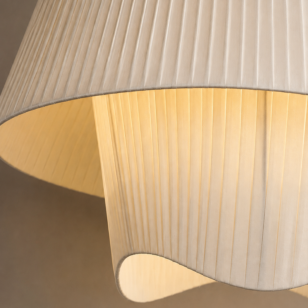 A close-up of a pleated fabric lampshade in a warm off-white or cream color, glowing with a soft warm light from within. The shade features vertical pleats and a curved, sculptural lower section.