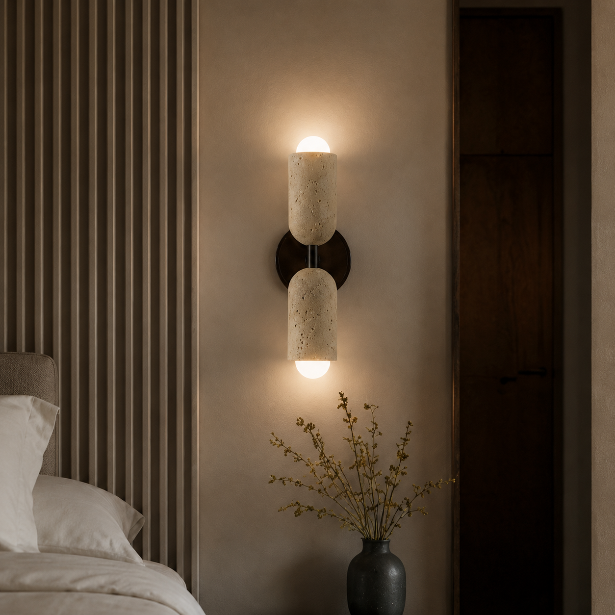 A modern wall-mounted double-light sconce with cylindrical textured stone shades and a dark metal base, illuminated in a bedroom setting next to a bed and a vase of dried branches.