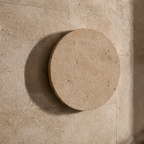 A round travertine stone pedestal or table top in a warm beige tone, shown from a top-down angle on a matching stone tiled floor.