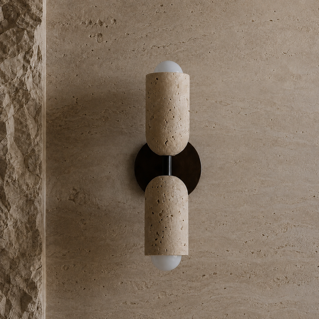 A modern wall-mounted sconce with two cylindrical beige stone shades and frosted white globe bulbs, mounted on a textured beige stone wall.