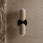 A modern wall-mounted sconce with two cylindrical beige stone shades and frosted white globe bulbs, mounted on a textured beige stone wall.