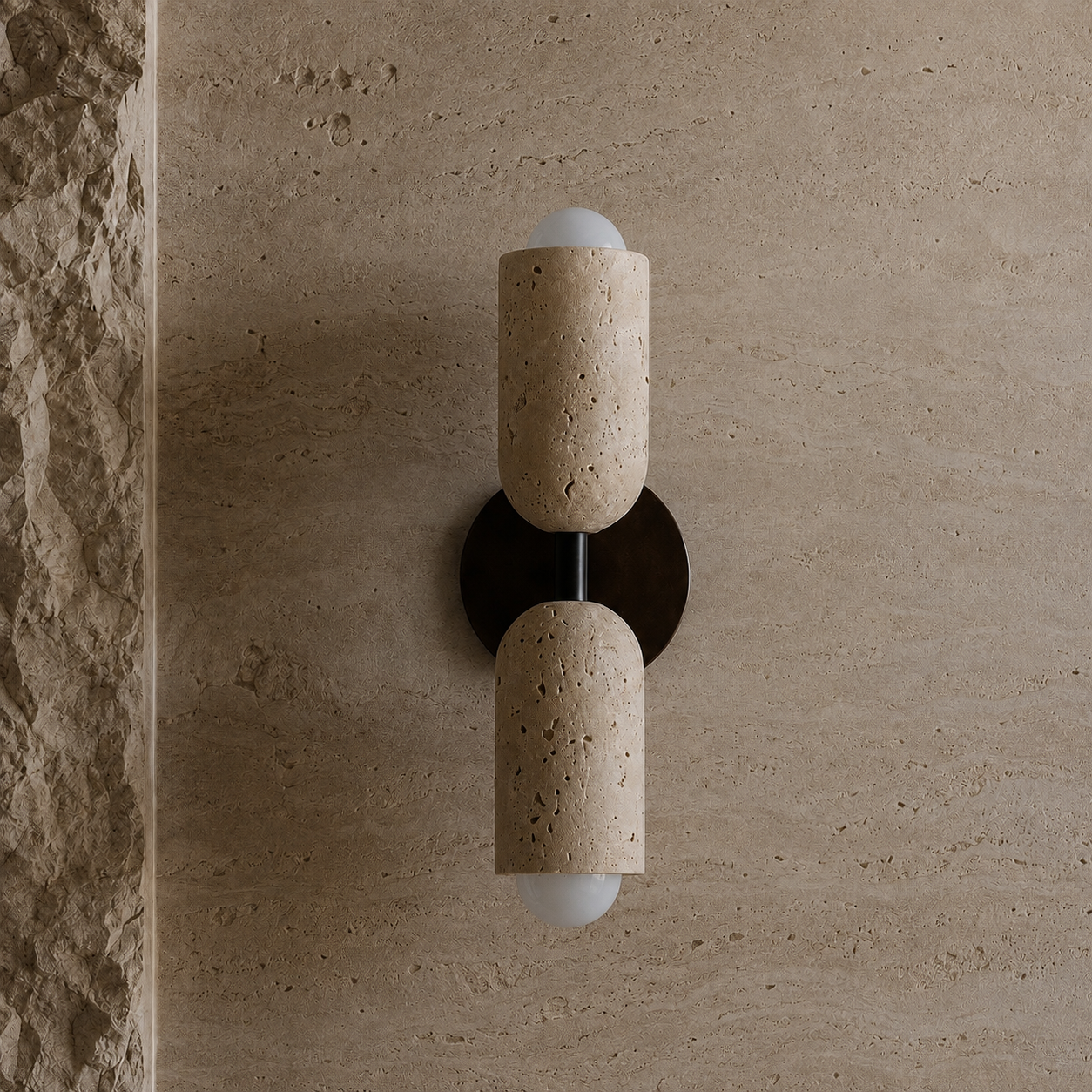 A modern wall-mounted sconce with two cylindrical beige stone shades and frosted white globe bulbs, mounted on a textured beige stone wall.