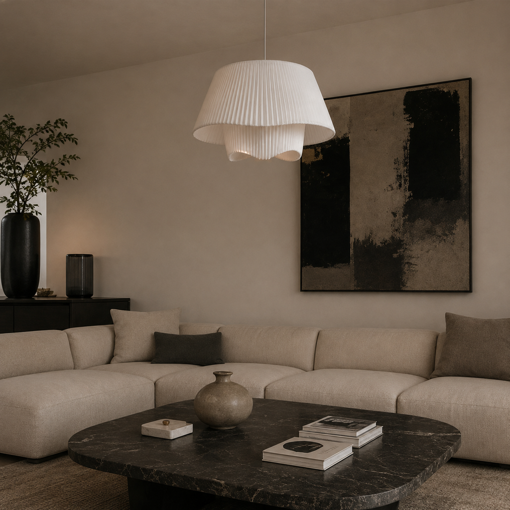 A modern living room scene featuring a large pleated white pendant light hanging over a beige sectional sofa. The room has warm neutral walls, a black-framed abstract art print, a dark marble coffee table, and a potted plant in a tall black vase.